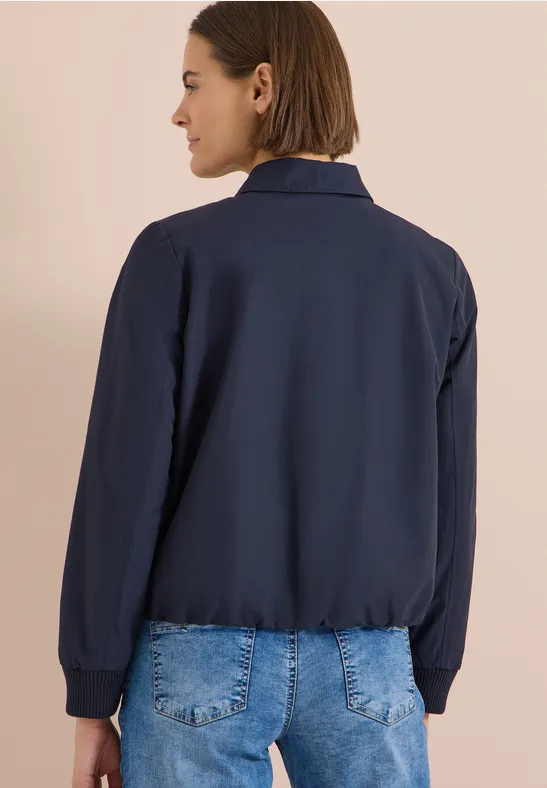 Rückansicht einer Person in dunkelblauer Jacke und blauer Jeans vor beigem Hintergrund.