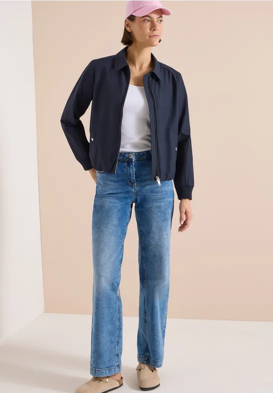 Frau in dunkelblauer Jacke, weißem T-Shirt und blauen Jeans vor hellem Hintergrund.