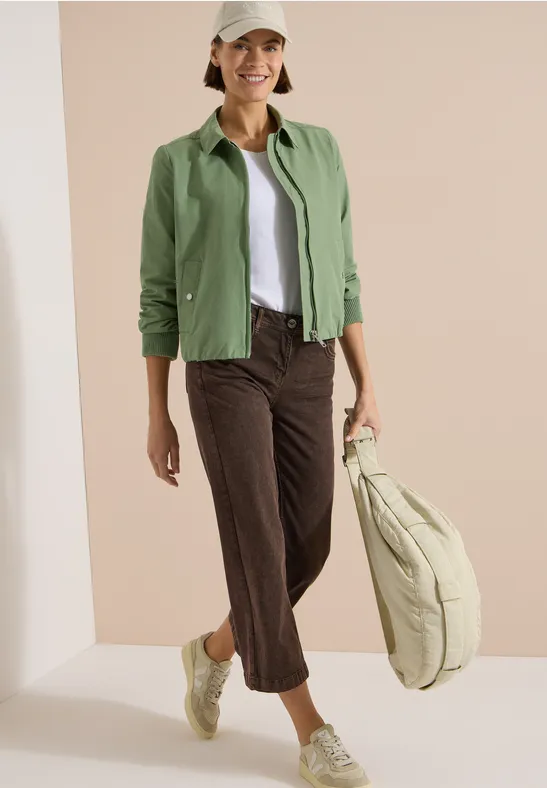 Femme en veste verte, top blanc, pantalon marron raccourci, baskets vertes et grand sac beige.