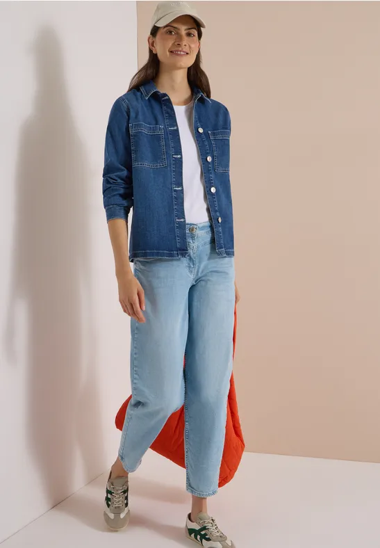 Frau in Jeansjacke, weißem T-Shirt, hellen Jeans und Sneakern trägt Cap und rote Tasche.