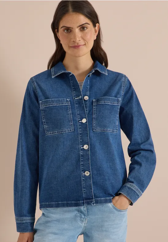 Frau in dunkelblauer Jeansbluse mit Brusttaschen und heller Jeans vor neutralem Hintergrund.