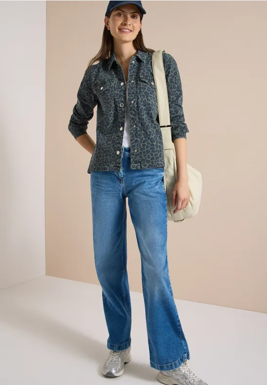 Frau in Jeans, gemusterte Bluse, Baseballkappe und Beuteltasche vor hellem Hintergrund.