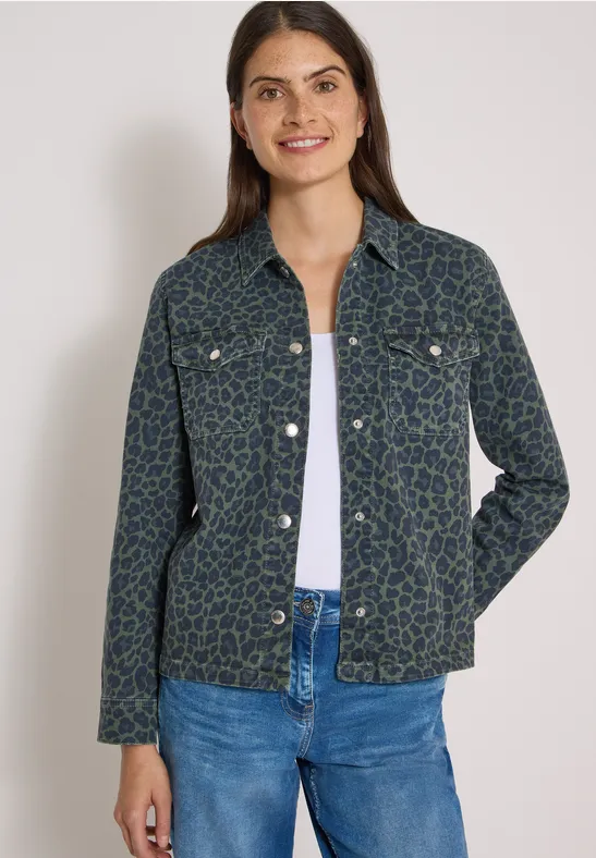 Frau in grüner Blouson-Jacke mit Muster, weißem Top und blauen Jeans vor hellem Hintergrund.