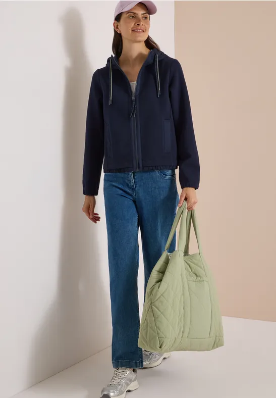 Een vrouw draagt een donkerblauwe hoodie, jeans, een pet en houdt een grote lichtgroene, gewatteerde tas vast.