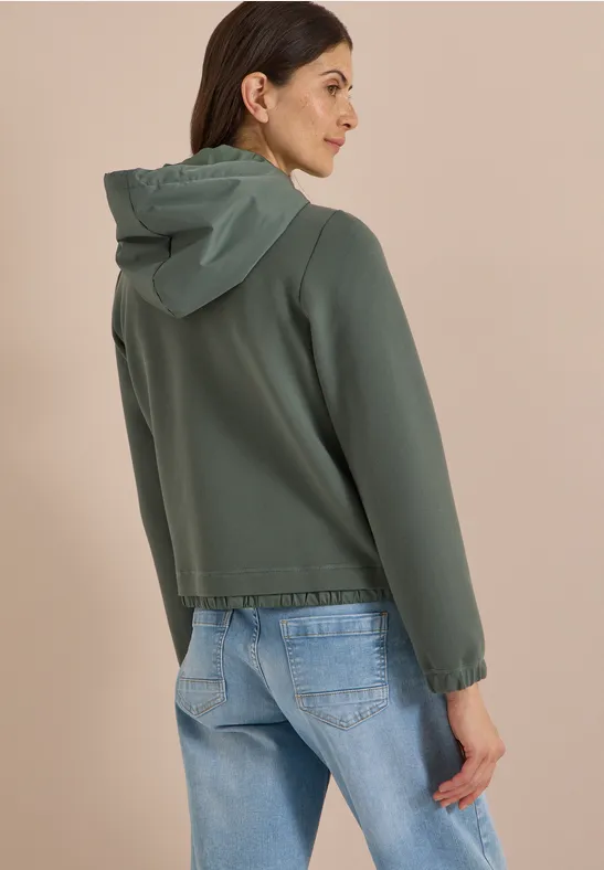 Eine Frau steht seitlich von hinten, trägt eine grüne Kapuzenjacke und blaue Jeans.