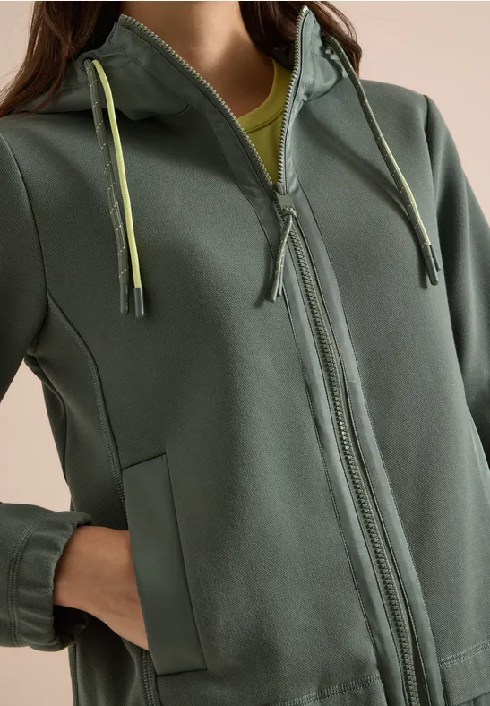 Person trägt graugrünen Kapuzenpullover mit Reißverschluss, seitlichen Taschen und hellgrünen Kordeln.
