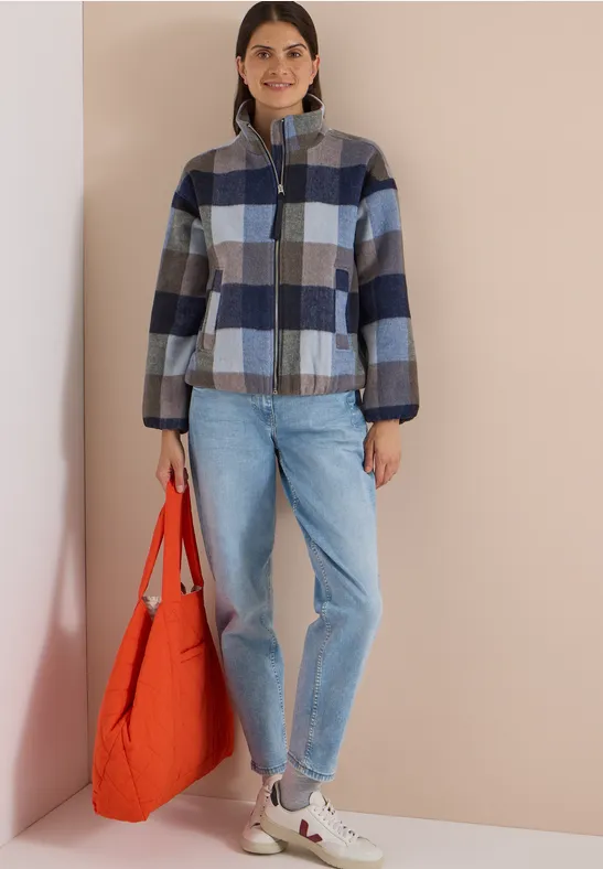Femme en veste à carreaux bleu-gris, jean et baskets blanches, tenant un sac orange.