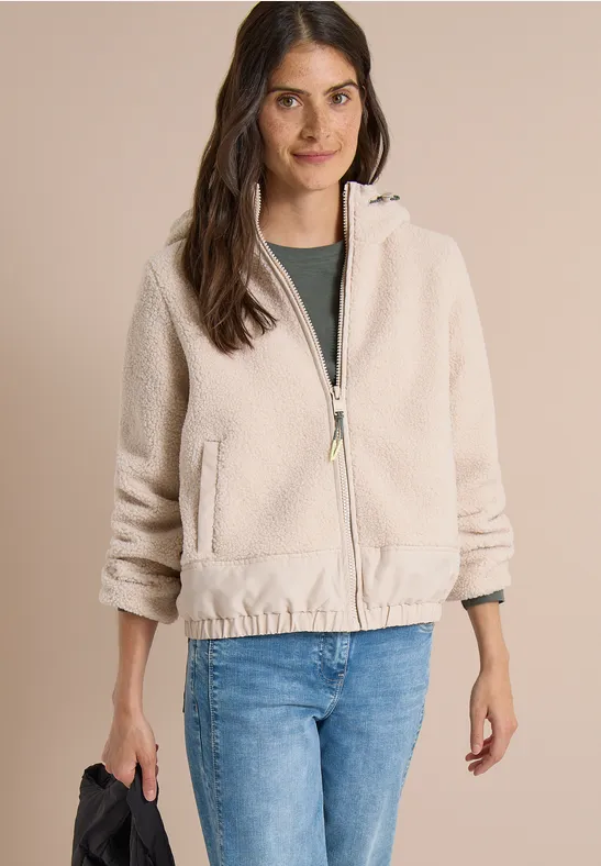Frau trägt eine beigefarbene Teddyjacke mit Reißverschluss und Taschen, kombiniert mit blauen Jeans.