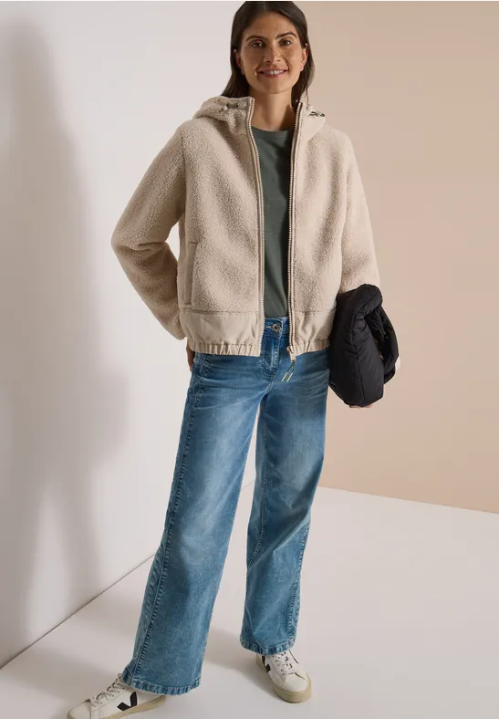 Frau in beiger Teddyjacke, grünem Shirt, blauen Jeans und weißen Sneakern, hält dunkle Jacke in der Hand.