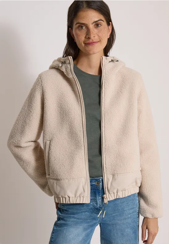 Frau in beige Sherpa-Fleecejacke mit Kapuze, grünem Shirt und Jeans, Hände in Taschen, lächelnd vor neutralem Hintergrund.