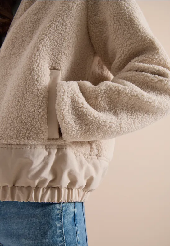 Beige Teddy-Fleecejacke mit elastischem Bund, Hand in Tasche gesteckt, kombiniert mit Jeans.