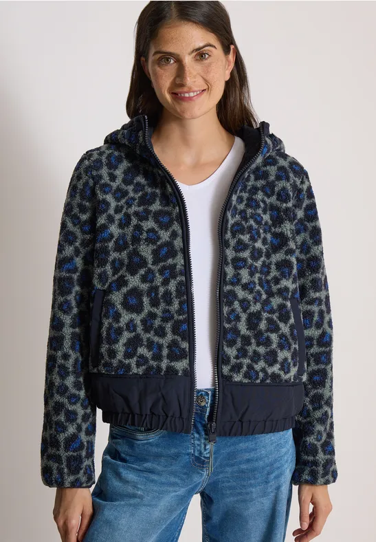 Een vrouw draagt een donkerblauwe capuchonvest met luipaardprint, een wit T-shirt en een blauwe jeans.