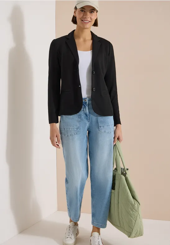 Frau in schwarzem Blazer, weißem Shirt, hellblauen Jeans und grüner gesteppter Tote Bag.