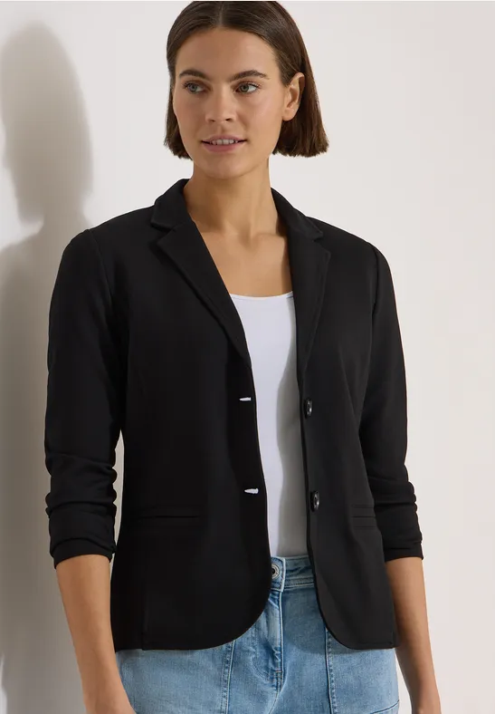 Frau in schwarzem Blazer, weißem Top und Jeans steht vor weißem Hintergrund.