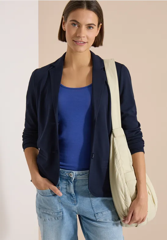 Frau in dunkelblauem Blazer, blauem Top und Jeans trägt eine beige Stepp-Tasche, lässig mit Hand in Tasche.