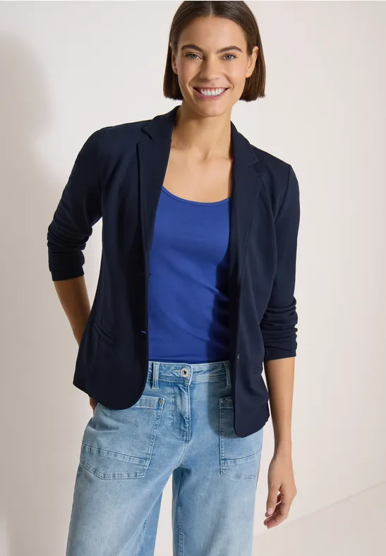 Frau mit dunkelblauem Blazer, blauem Top und hellblauen Jeans vor hellem Hintergrund.