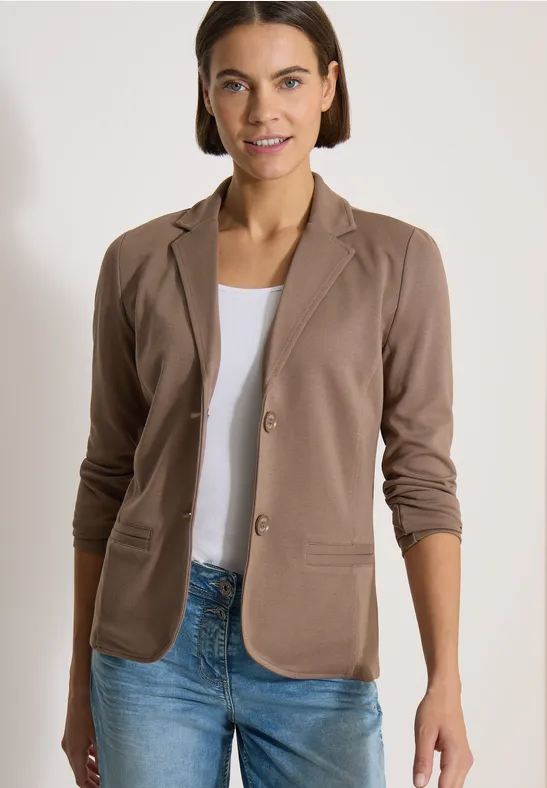 Frau in braunem Blazer, weißem Shirt und Jeans, stehend vor neutralem Hintergrund.