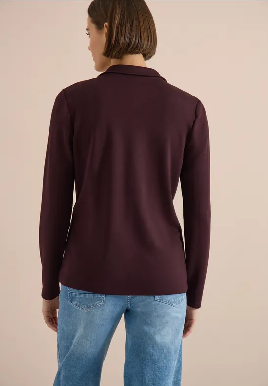 Frau von hinten in dunkelviolettem Langarm-Polo und blauen Jeans vor neutralem Hintergrund.