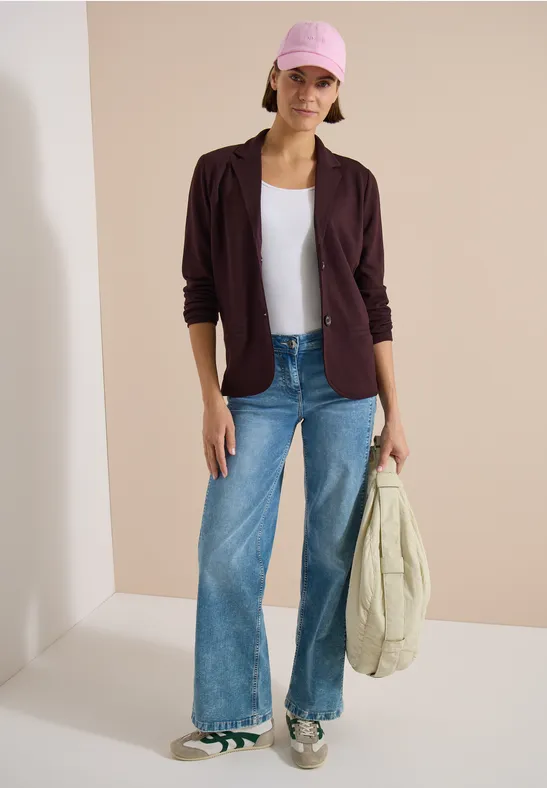 Eine Person steht in weitem Jeans, weißem Shirt, dunkler Jacke und Sneakers und hält eine helle Tasche.