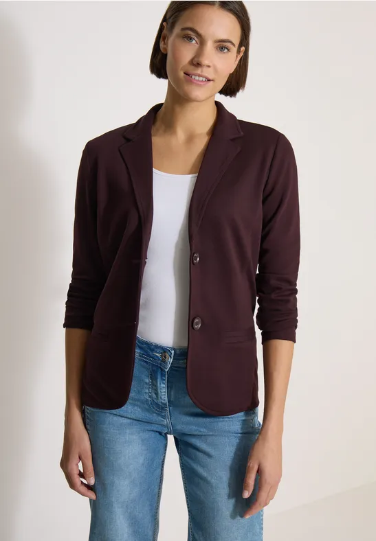 Frau im bordeauxroten Blazer mit weißem Top und blauen Jeans vor neutralem Hintergrund.