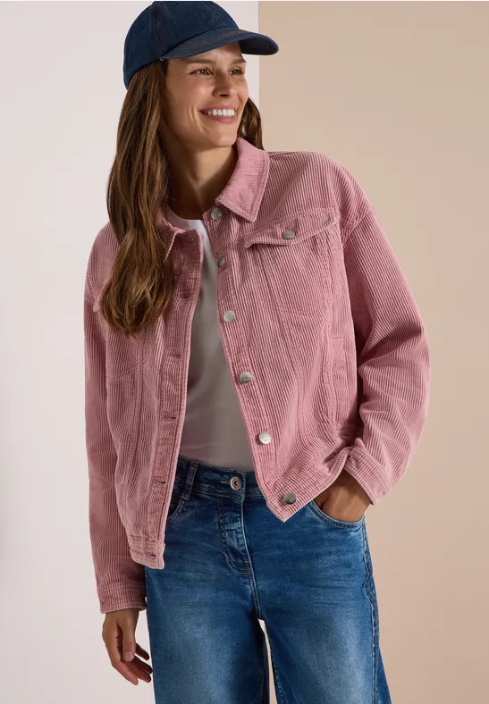 Jeune femme souriante en veste rose en velours côtelé, jean et casquette bleue.