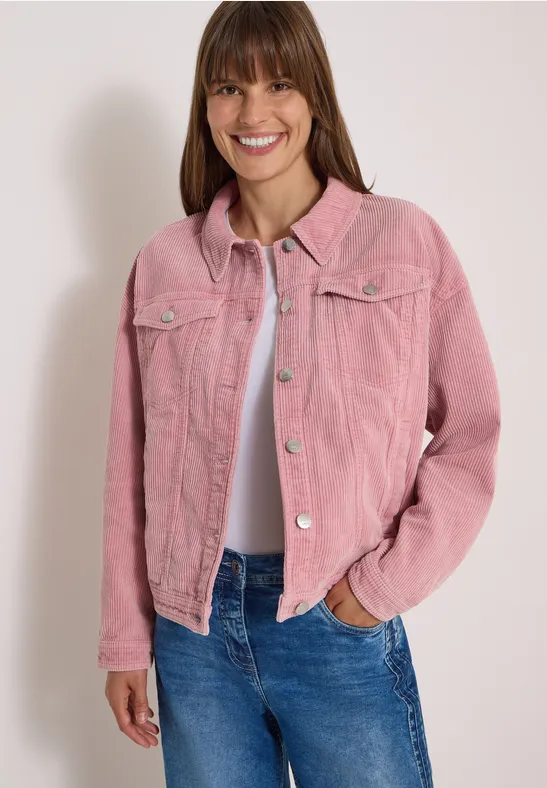Femme souriante en veste rose en velours côtelé, t-shirt blanc et jean bleu, fond neutre.