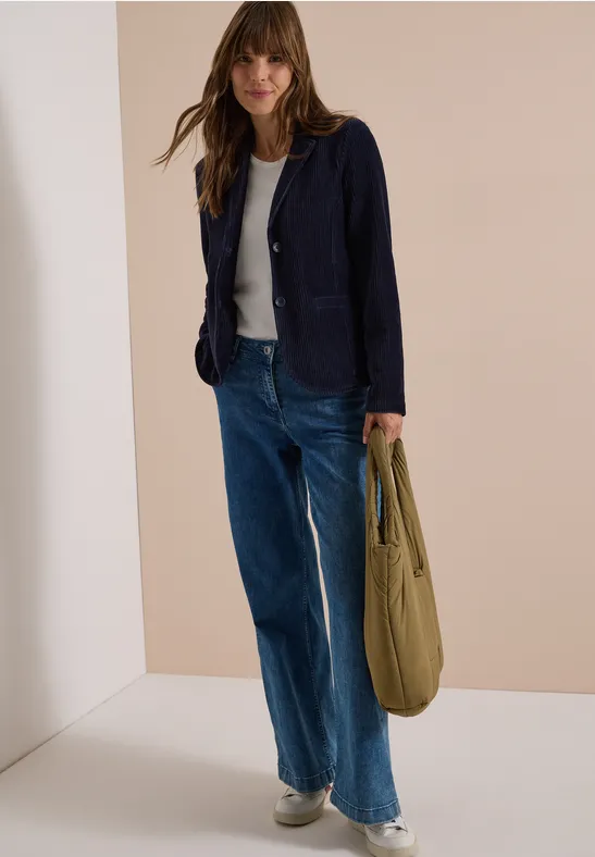 Frau in weiter Jeans, weißem T-Shirt und dunkelblauem Blazer mit olivgrüner Tasche.