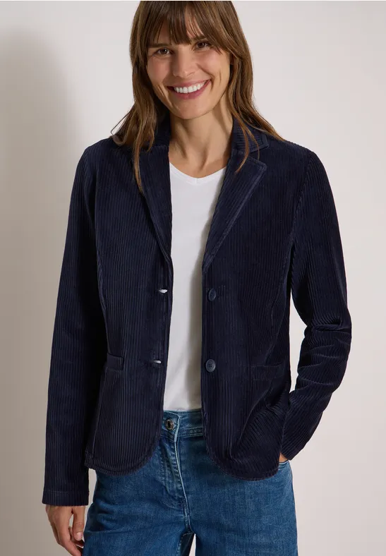 Frau in dunkelblauem Blazer, weißem T-Shirt und Jeans, lächelnd.