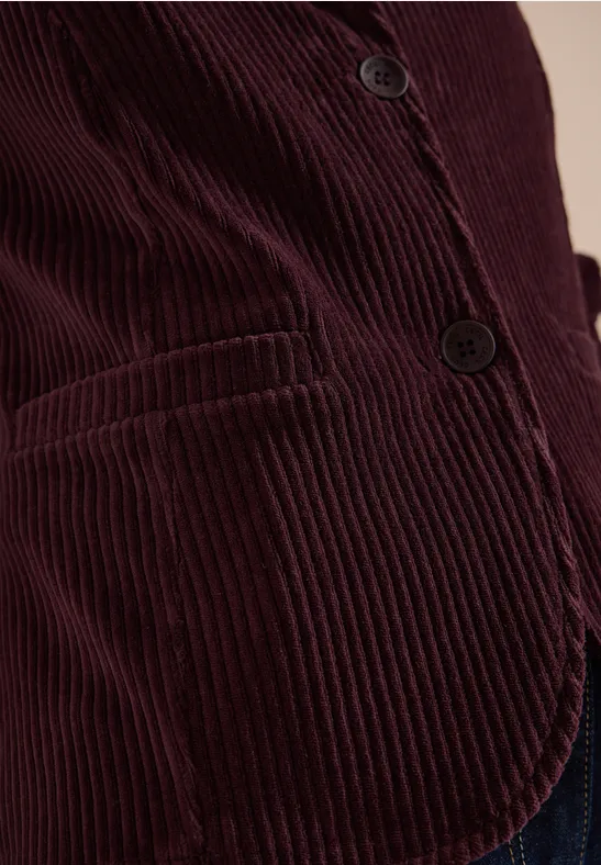 Nahaufnahme einer Person in einer burgunderroten Cord-Feinrippjacke mit Knöpfen und Tasche.
