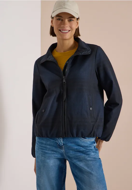 Frau in dunkelblauer Jacke, Jeans und Basecap vor hellem Hintergrund, lächelnd und entspannt.