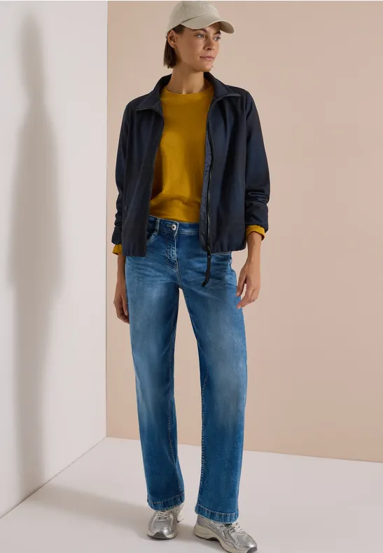 Frau trägt blaue Jeans, senfgelber Pullover, dunkelblaue Jacke und beige Mütze vor hellem Hintergrund.