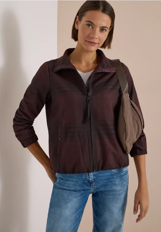 Frau in legerer Jacke, Jeans und Schultertasche lehnt entspannt an einer weißen Wand.
