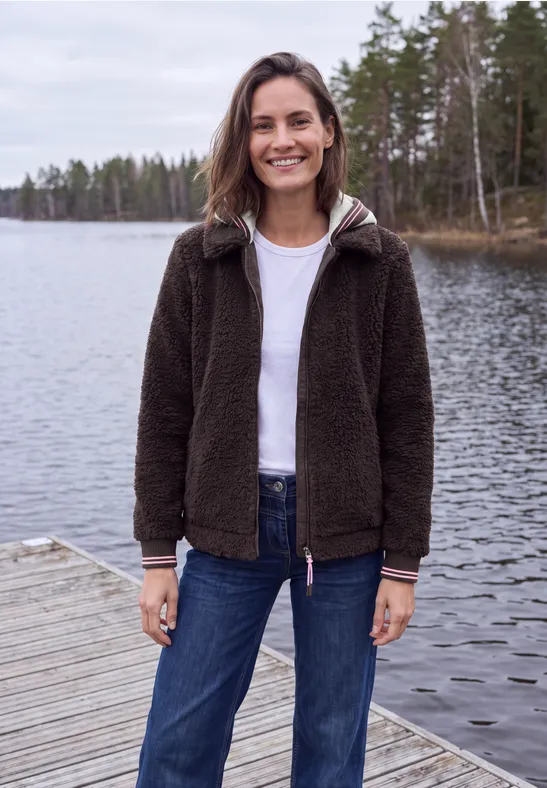 Frau lächelt auf Steg am ruhigen See, trägt dunkle Jacke, weißes Shirt und Jeans vor Waldkulisse.