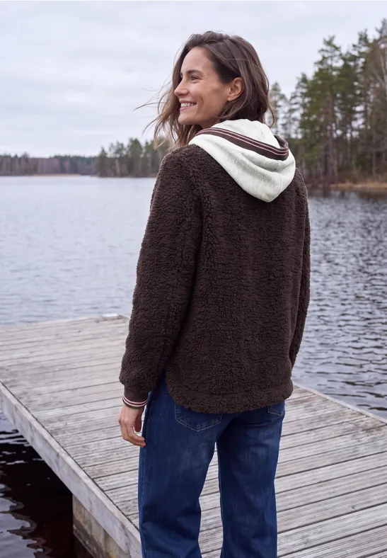 Frau in braunem Fleece-Hoodie steht lächelnd auf einem Steg am See, umgeben von Bäumen und Wasser.