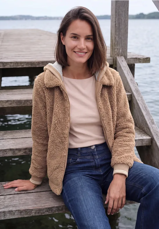 Junge Frau sitzt auf einer Holztreppe am Wasser, lächelt und trägt Jeans und eine kuschelige Jacke.