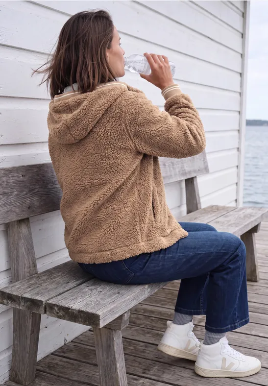 Frau in brauner Teddyjacke und Jeans sitzt auf einer Holzbank vor weißer Hütte am Wasser und trinkt.