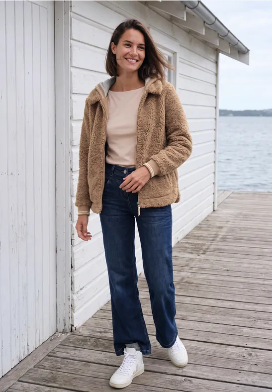 Eine Frau in heller Teddyjacke, Jeans und weißen Sneakern steht lächelnd an einer weißen Holzlaube am See.
