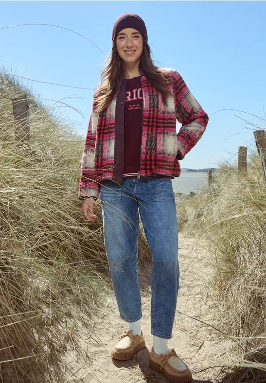 Frau in kariertem Hemd und Jeans mit Mütze steht auf Dünenpfad am Strand bei klarem Himmel.
