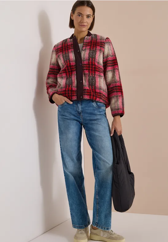 Frau vor weißer Wand trägt rote-pinke Karo-Jacke, blaue Jeans und hält eine schwarze Tasche.