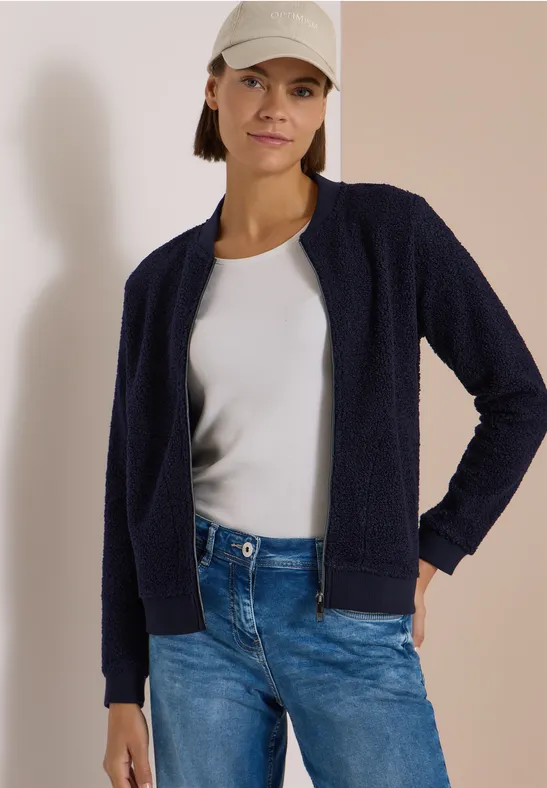 Frau mit dunkelblauer Strickjacke, weißem Shirt, Jeans und beiger Baseballkappe.