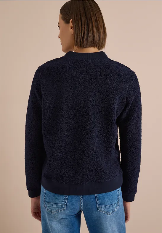 Eine Person von hinten trägt einen dunkelblauen Teddy-Pullover vor beigem Hintergrund.