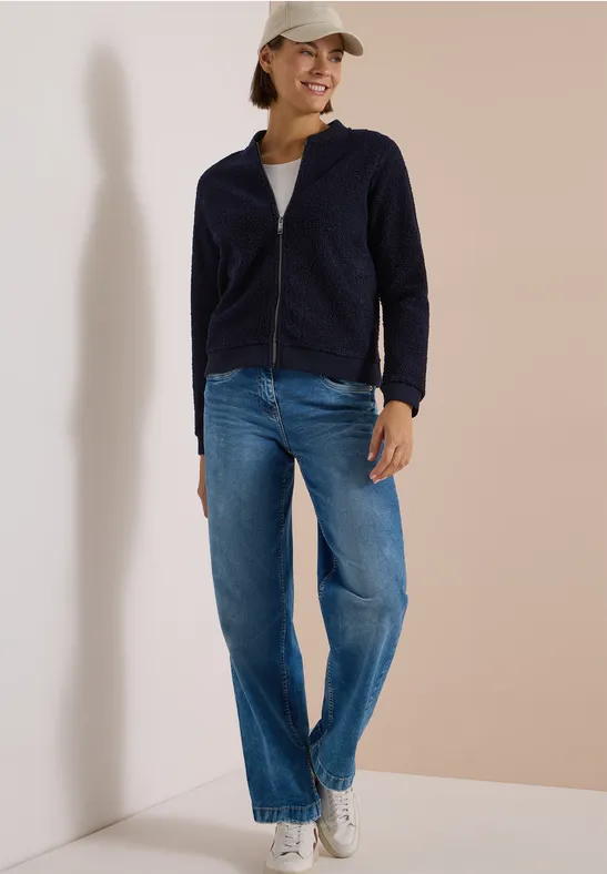 Frau mit blauer Jacke, Jeans und Cap lehnt lässig an eine helle Wand.