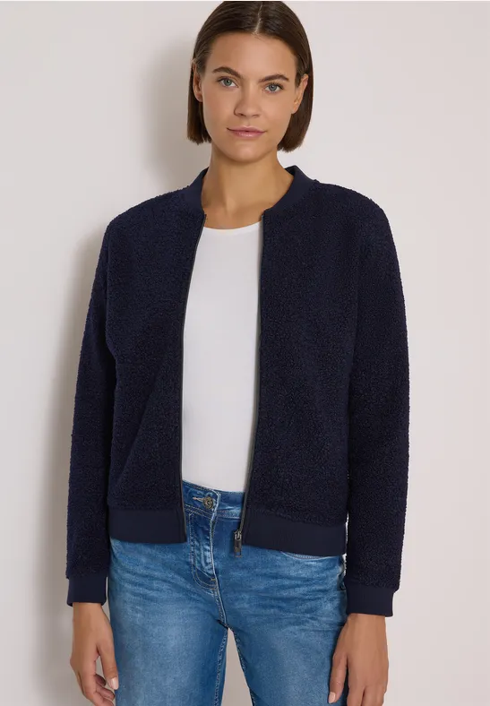 Frau mit kurzen braunen Haaren trägt dunkelblaue Fleece-Jacke, weißes Shirt und blaue Jeans.