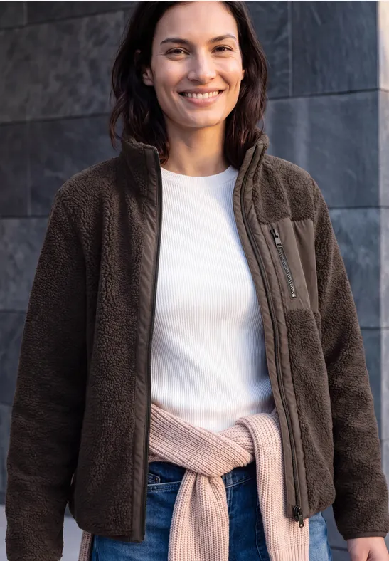 Een glimlachende vrouw draagt een bruine fleecejas, een wit shirt en heeft een vest om haar middel geknoopt.