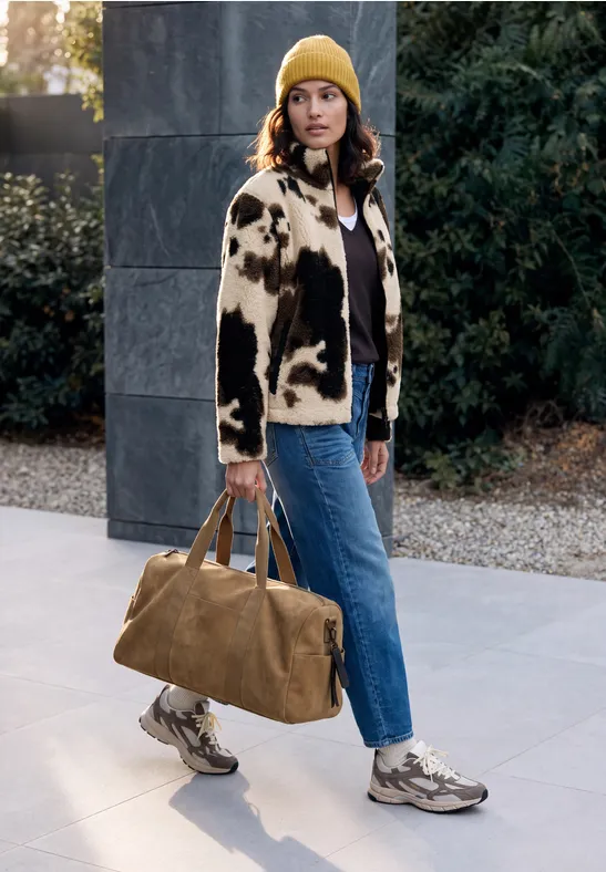 Eine Frau in modischem Freizeitlook mit Jeans, Felljacke, gelber Mütze und Sneakern trägt eine Reisetasche.