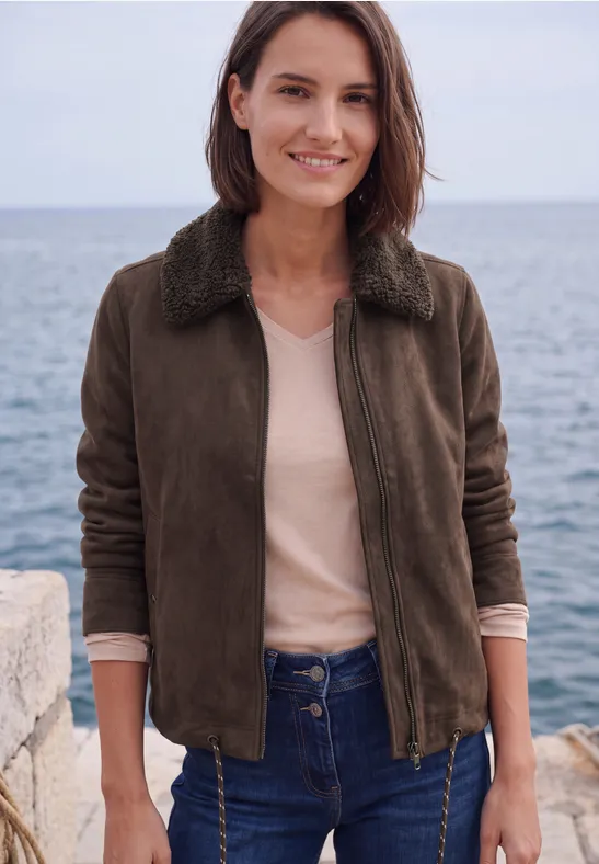 Frau im braunen Blouson vor dem Meer, lächelt, trägt Jeans und beiges Shirt.