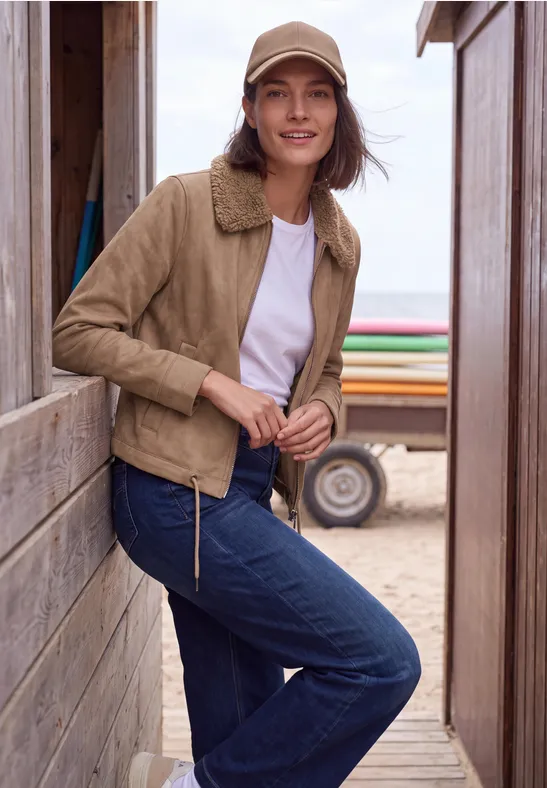 Frau in beiger Jacke, Jeans und Cap lehnt an Holzwand am Strand, im Hintergrund Sand und Meer.