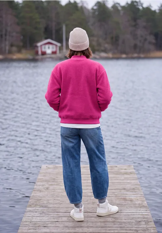 Persoon in roze trui en witte muts staat op steiger, kijkt uit over rustig meer met huisje in bos.