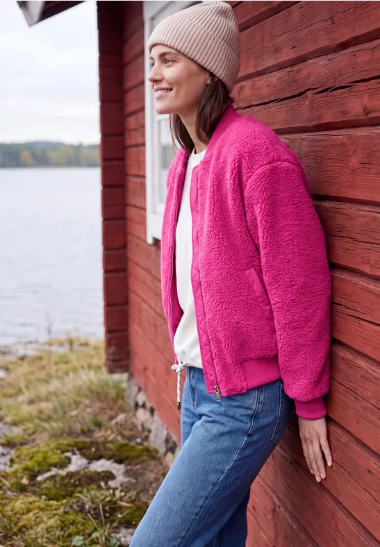 Een vrouw in een roze fleecejas leunt glimlachend tegen een rood houten huisje bij het meer.