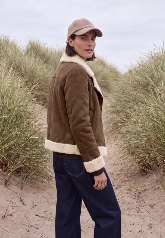 Eine Frau in brauner Lammfelljacke, Jeans und Basecap steht in Dünen mit Gräsern am Strand.